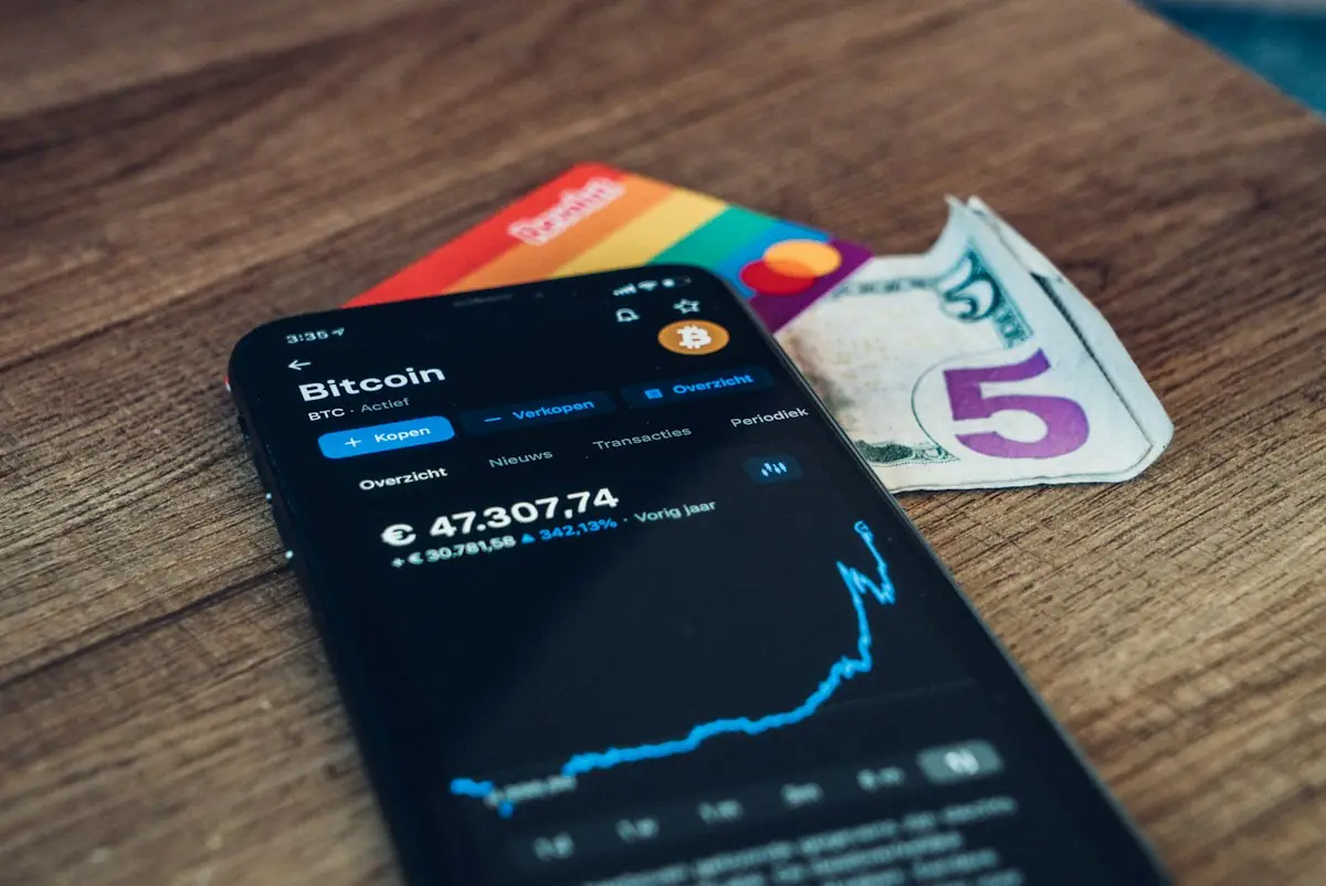 A crypto app showing a bitcoin price on a phone beside cards and cash